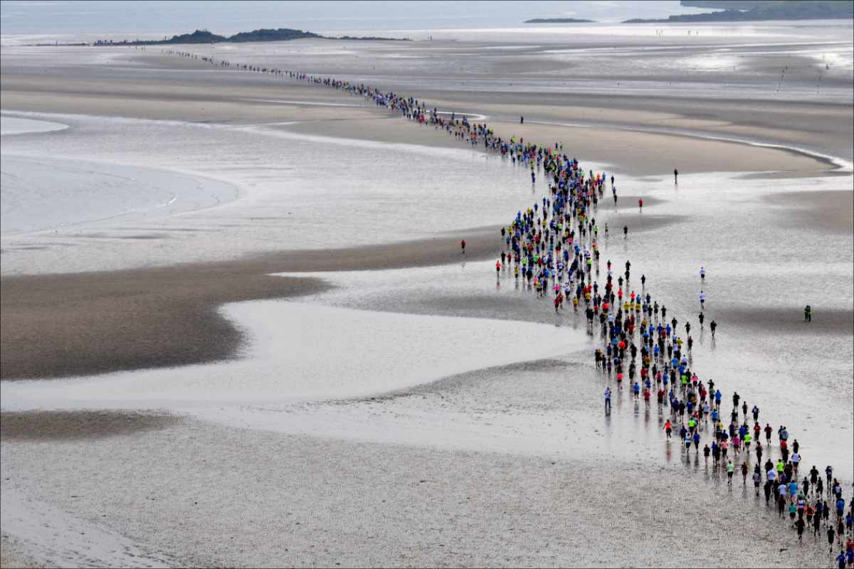 photo of a stream of runners crossing wet sand with rocks and sea in the background.